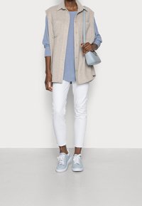 Person wearing a beige sleeveless buttoned vest, blue long-sleeve top, white cropped jeans, light blue sneakers, and carrying a light blue shoulder bag.