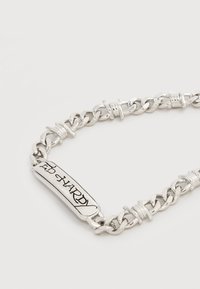 Ασημένιο βραχιόλι με αλυσίδα, με σύνδεσμο σε σχήμα ράβδου που έχει χαραγμένο το "ED HARDY" με στυλιζαρισμένη μαύρη γραφή σε λευκό φόντο.