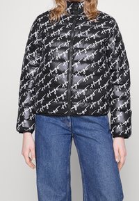 Puffjakke i blank sort med hvidt skrifttekst. Har en høj krave og lynlås. Bæres med blå vide jeans.