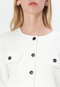 Weißer strukturierter Blazer mit rundem Halsausschnitt, vier Taschen und dekorativen schwarzen Knöpfen. Hat ein dezentes gewebtes Muster.