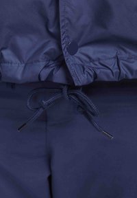 Marinblå vattentät jacka med justerbar dragsko, tryckknappslås i nederkant och slät yta. Har elastik i midjan för justering av passformen.