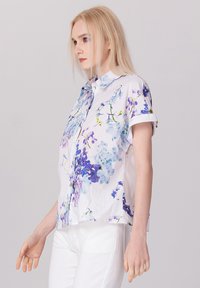Chemise à manches courtes avec boutons, de couleur blanche, ornée d'un motif floral dans des teintes de bleu et de violet. Fabriquée en tissu léger avec une coupe décontractée.