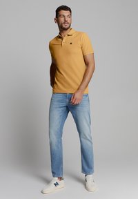 Korte mouwen polo in mosterdgeel, met een puntkraag, een placket met twee knopen en een klein logo. Gekoeld met lichtblauwe jeans en sneakers.