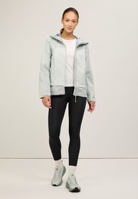 Ljusgrön vattentät jacka med huva, meshpaneler, dragkedjeslutning, kombinerad med svarta leggings och ljusgröna träningsskor.