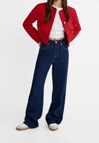 Rode cropped jas met rafelige randen, gecombineerd met wijde donkerblauwe jeans. Witte t-shirt eronder en beige sneakers maken de outfit compleet.