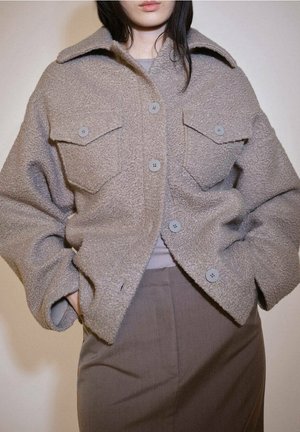 Mujer con chaqueta de lana texturizada color topo abotonada con grandes bolsillos en el pecho sobre top gris y pantalones marrones de talle alto, manos en los bolsillos.
