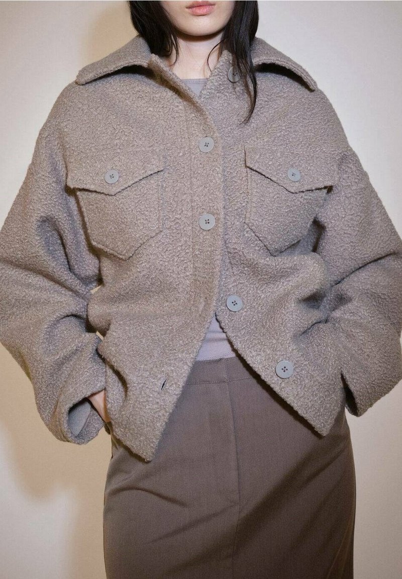Femme portant une veste en laine texturée taupe boutonnée avec de grandes poches poitrine sur un haut gris et un pantalon taille haute marron, mains dans les poches.