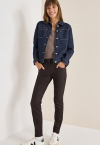 Džínová bunda v tmavě modré barvě s dvěma předními kapsami, zapínání na knoflíky a béžovým topem. Model má na sobě hnědé kalhoty a bílé tenisky s černými detaily.
