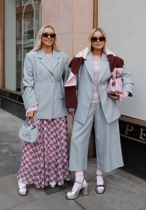 Zwei Frauen, die graue Blazer und Sonnenbrillen tragen; eine mit gemustertem Rock, die andere mit verkürzten Hosen, pinkfarbenen Accessoires und silbernen Absätzen.