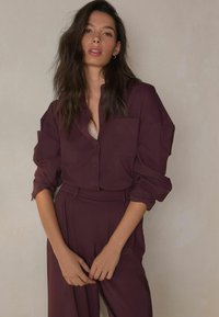 Burgundy shirt with button closure and chest pocket, paired with matching loose trousers; both feature soft, flowing material and relaxed fit.