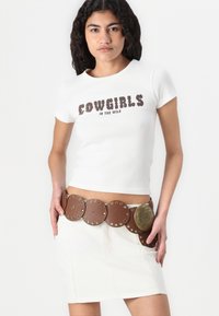 Balts T-krekls ar uzrakstu "COWGIRLS IN THE WILD" brūnā krāsā. Brūns ādas jostas, dekorēta ar apaļām akcentiem un metāla detaļu, uzvilkta virs balta svārka.