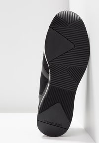 Semelle de chaussure de sport noire en caoutchouc texturé, présentant des rainures et des accents lisses. "MICHAEL KORS" embossé sur la zone du talon.