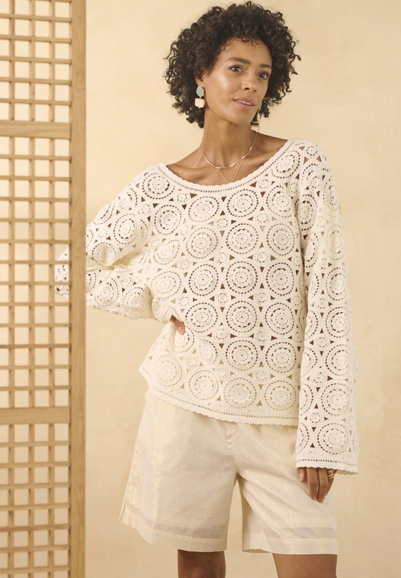 Witte haakpullover met cirkelpatronen en brede mouwen, gedragen met crèmekleurige linnen shorts. Eenvoudige sieradenaccenten maken de look compleet.