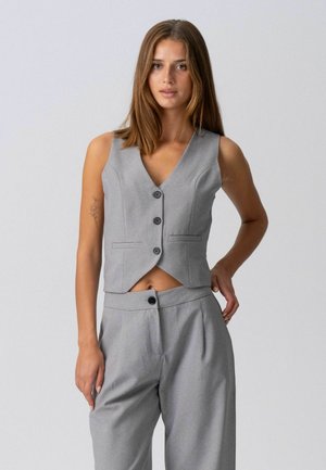 Sirgetepruunide juustega naine, kandes halli varrukateta nööpidega vest ja sobivaid kõrge vöökohaga pükse, seisab ühevärvilise tausta ees.