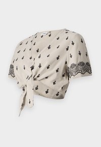 Blouse courte beige avec motifs géométriques noirs, manches courtes, devant noué et encolure en V profonde. Tissu texturé avec accents brodés.