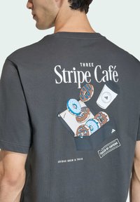 Šedé bavlněné tričko s grafickým designem donutů a kávy, text "THREE Stripe Café," s hladkou texturou a volným střihem.