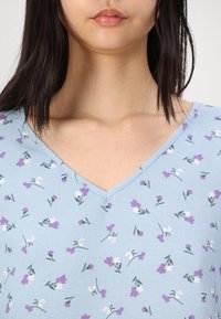 Top floral azul con diseño de escote en V, hecho de tejido texturizado, adornado con pequeños patrones de flores púrpuras y blancas.