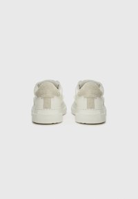 Sneaker bianche con tomaia in pelle liscia, dettagli in suede grigio sul tallone e suola in gomma texturizzata. La vista posteriore mostra i dettagli del collare imbottito.