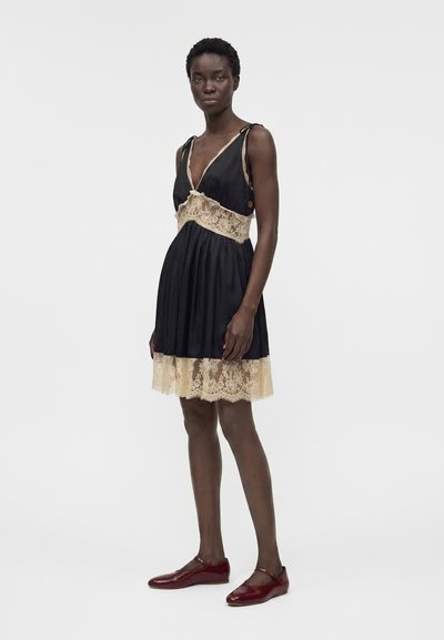 Γυναίκα με μαύρο αμάνικο φόρεμα με μπεζ δαντελένια λεπτομέρεια και κόκκινα παπούτσια Mary Jane, στέκεται μπροστά σε απλό ανοιχτόχρωμο φόντο.