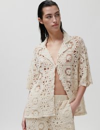 Femme portant une chemise en crochet beige et un short assorti, une main dans la poche, regardant sur le côté devant un fond uni.