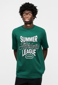 Πράσινο βαμβακερό T-shirt με μεγάλο λευκό κείμενο που αναγράφει "ΘΕΡΙΝΗ ΑΘΛΗΤΙΚΗ ΛΙΓΚΑ" και λογότυπο "adidas". Κοντά μανίκια και σχέδιο με στρογγυλή λαιμόκοψη.