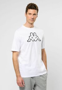 Fehér, kerek nyakú póló, amely pamutból készült, a mellkasán egy nagy fekete Kappa logóval. A modell szürke melegítőnadrággal párosítja.