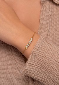 Pulsera de cadena de oro con cuentas multicolores y acentos hexagonales, llevada en una muñeca con un fondo de tela texturizada de color marrón claro.