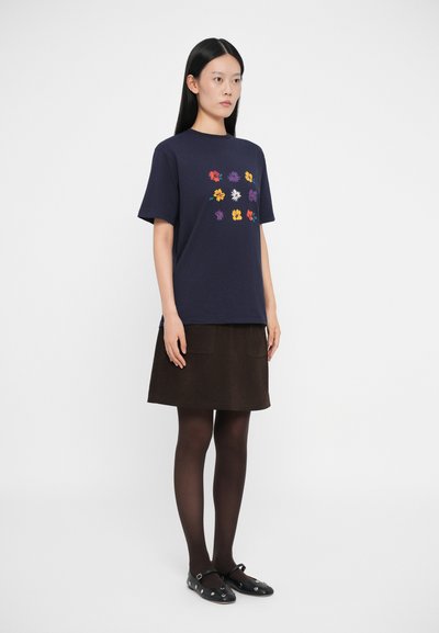 Camiseta de algodón azul marino con mangas cortas que presenta un colorido bordado floral en la parte frontal. Combinada con una falda marrón y medias oscuras.