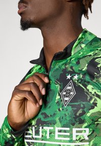 Grøn langærmet sports skjorte med sort krave, der har camouflage mønster, logo og fed hvid tekst på tværs af fronten.