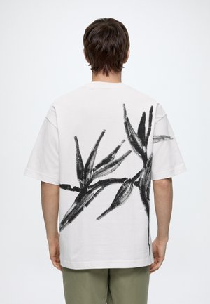 Mand iført hvid t-shirt med stort sort abstrakt plante-design på ryggen, kombineret med olivengrønne bukser, stående med ryggen mod.