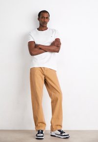 Camicia bianca a maniche corte con bordi rossi, pantaloni beige a gamba dritta e sneaker bianche e nere. Design minimalista su uno sfondo neutro.