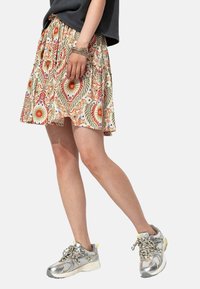 Jupe mi-longue à motif floral avec une base beige, ornée de détails rouges, verts et bleus ; associée à des baskets en métal argenté.