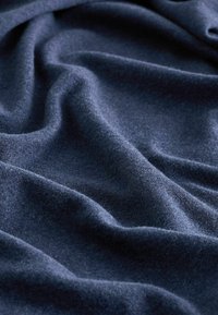 Jūrinės mėlynos spalvos audinys su minkšta, tekstūruota paviršiumi, kuriame matyti raukšlės ir drapiruotės, pabrėžiančios jo storį ir pūkuotą kokybę.