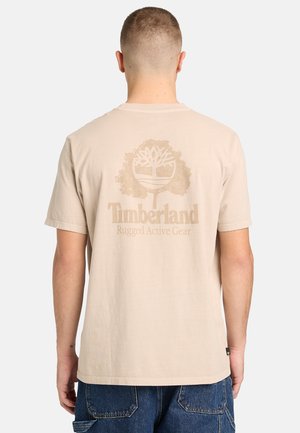 Homme vu de dos portant un t-shirt beige Timberland avec un logo d'arbre et le texte "Rugged Active Gear", associé à un jean en denim bleu.