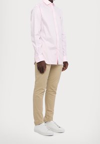 Světle růžová košile s dlouhým rukávem a límečkem, kombinovaná s béžovými slim-fit kalhotami a bílými teniskami. Jsou zde patrné horizontální pruhy.