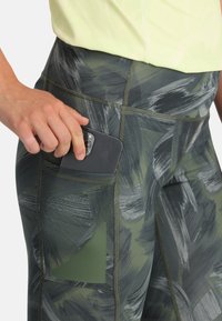 Una mano inserisce uno smartphone nero nella tasca laterale di leggings sportivi verde e grigio a motivo, indossati con una maglia gialla chiara.