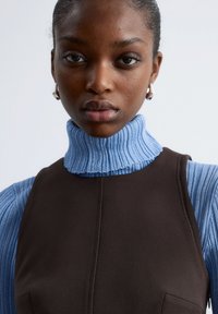 Gestufte Outfit mit einem gerippten hellblauen Rollkragenpullover und einem dunkelbraunen ärmellosen Kleid, mit sichtbarer Naht und einem seitlichen Taschen-Detail.