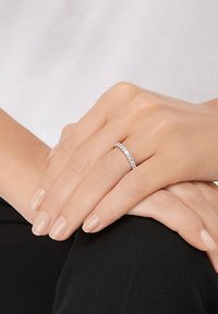 Bague en argent ornée de diamants avec un anneau courb�é, mettant en valeur une série de petites pierres précieuses transparentes. Des mains reposent calmement sur un genou.