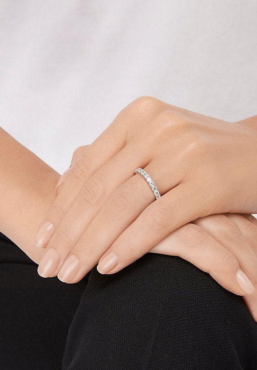 Bague en argent ornée de diamants avec un anneau courbé, mettant en valeur une série de petites pierres précieuses transparentes. Des mains reposent calmement sur un genou.