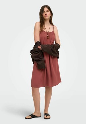 Jeune femme portant une robe sans manches rose poudrée, des sandales noires, un collier pendentif, et tenant une veste sombre autour de ses bras.