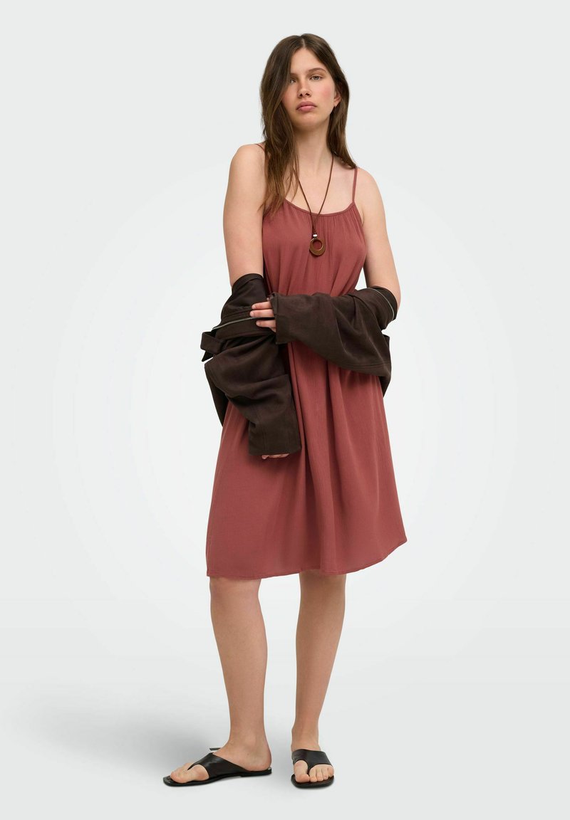 Jeune femme portant une robe sans manches rose poudrée, des sandales noires, un collier pendentif, et tenant une veste sombre autour de ses bras.