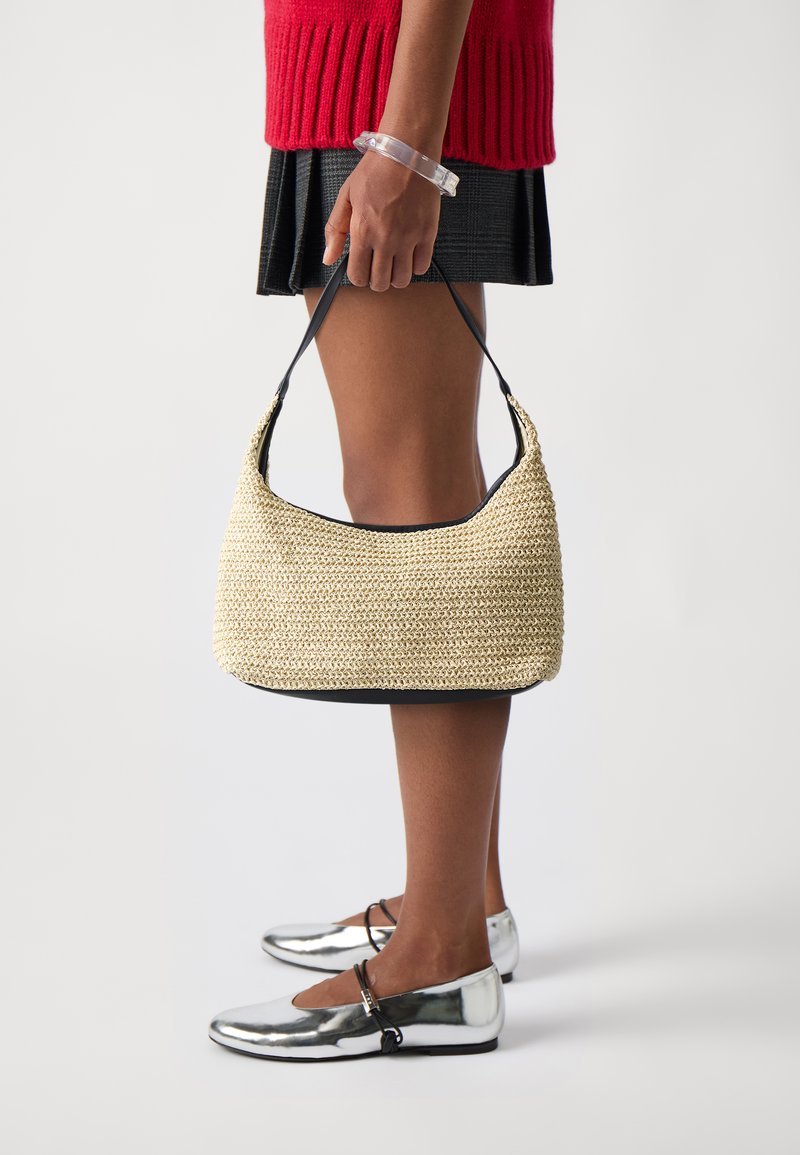 Sac à main en osier beige avec une forme courbée et des finitions en cuir noir. Le mannequin le tient avec une seule anse. Chaussures plates argentées brillantes avec une lanière.