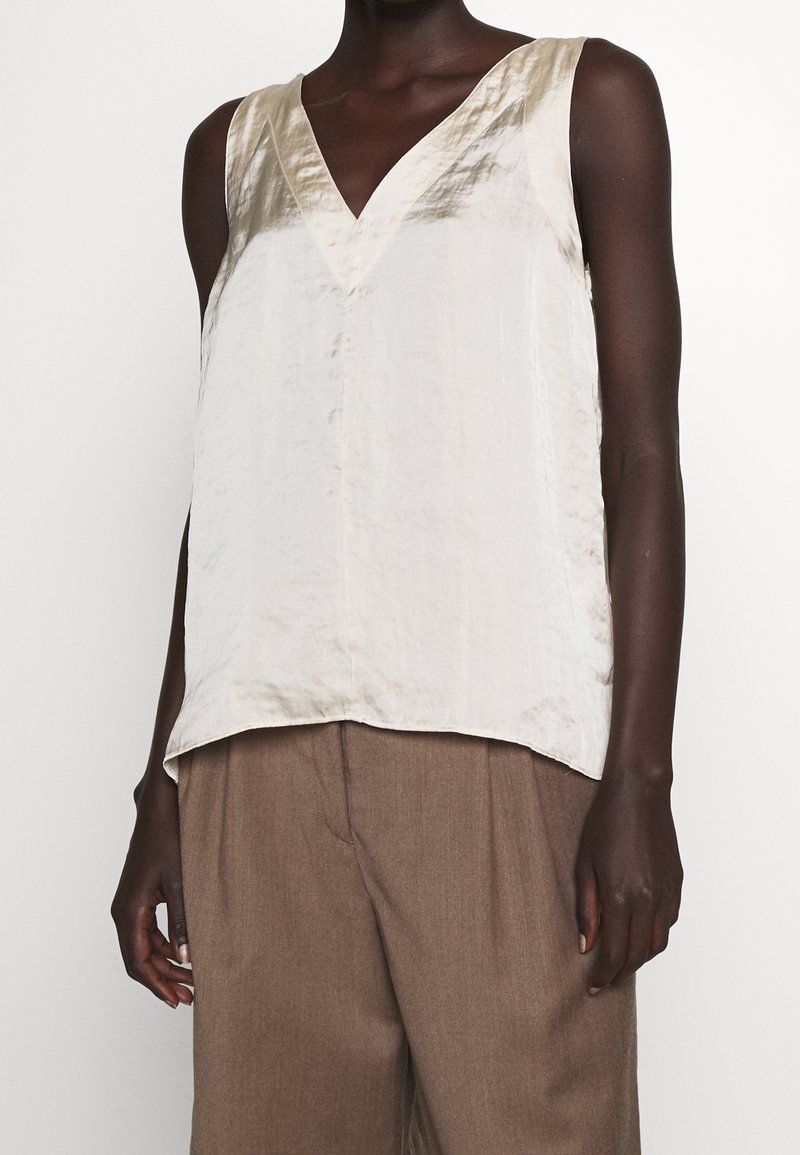 Top senza maniche in seta color crema con scollatura a V e spacchi laterali. Texture liscia e vestibilità leggermente ampia, indossato con pantaloni a vita alta marroni.