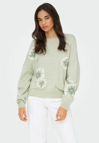 Pull en maille vert clair orné de motifs floraux blancs et verts, avec des épaules tombantes et un ourlet côtelé, associé à un pantalon blanc.