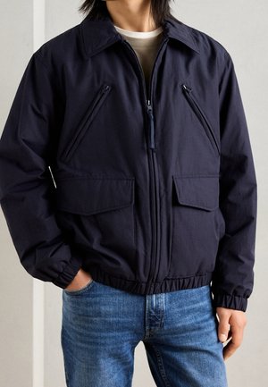 Person iført en mørk marineblå lynlåstrøje med skrå lynlåslommer på brystet og klaplommer i taljen, kombineret med blå jeans.