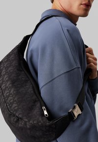 Bolso cruzado negro con un patrón sutil, cierre de cremallera y herrajes metálicos, llevado sobre un sudadera azul, que muestra un diseño elegante.
