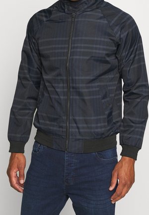 Mann trägt eine dunkelblaue Karierten Reißverschlussjacke mit gerippten Bündchen und Saum, kombiniert mit dunkelblauen Jeans, steht vor einfachem Hintergrund.