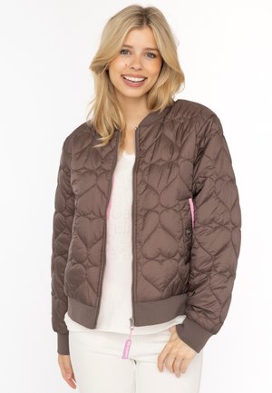Lächelnde Frau mit blonden Haaren trägt eine braune gesteppte Jacke mit Herzsteppung, ein weißes Hemd und weiße Hosen vor weißem Hintergrund.