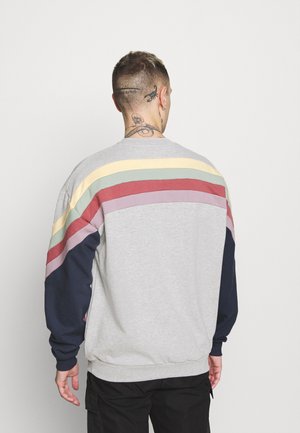Persoon met kort haar en meerdere tatoeages op de nek en het hoofd, gekleed in een grijze trui met pastelkleurige regenboogstrepen en zwarte broek, van achteren gezien.