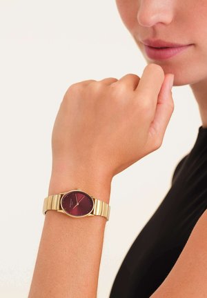 Montre dorée avec un cadran rond bordeaux et un bracelet en maillons, présentant une finition lisse et un design minimaliste.